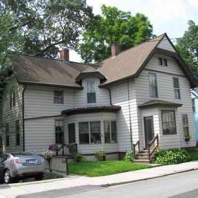 176 Prospect Street, Willimantic, CT, from the 2024-25 Tax Assessor's Database.: The image shows a two-story house with a traditional design, featuring a light beige exterior adorned with dark trim that contrasts the subtle tones. The roof, with its steep pitch, is covered in brown shingles, adding both durability and aesthetic appeal. Large windows punctuate the facade, allowing ample natural light to flood the interior spaces. A short set of steps leads to the main entrance, which is flanked by neatly trimmed shrubs and small patches of green grass, contributing to a welcoming ambiance. To the side, a grey car is parked on the driveway, which runs parallel to a concrete sidewalk, suggesting a quiet suburban neighborhood setting. A wooden railing protects the porch area, decorated with an array of potted plants.