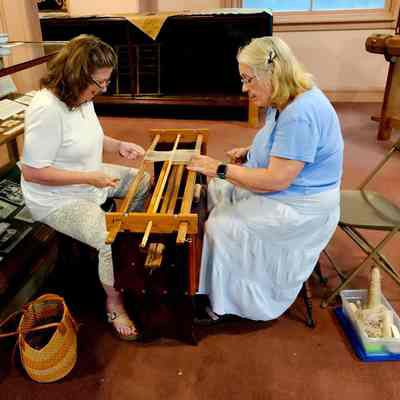 Volunteer weavers Susan Bruno (L) and Peggy Church (R) constructing the new string heddles.: In the image, two women are sitting opposite each other, deeply focused on working with a wooden device for making string heddles, a traditional piece of equipment used for textile creation, situated in a room that appears to be part of a historical or educational exhibit. One woman, clad in a white top and patterned pants, attentively adjusts the strings, while the other, wearing a blue top and light-colored skirt, mirrors her concentration. The setting suggests a vintage or museum-like environment, with glass display cases showcasing historical documents and artifacts, while a woven basket and container filled with yarn and tools are positioned nearby, hinting at a demonstration or workshop aimed at preserving or teaching historical textile-making techniques. The scene reflects a blend of tradition and community engagement.