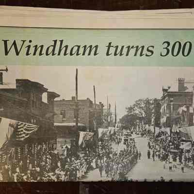 Cover of 300th Anniversary of Windham Commemorative Edition of the Willimantic Chronicle: The image shows a newspaper with the headline "Windham turns 300," featuring a historical photograph of a lively street scene, likely celebrating the town's tricentennial. The street is lined with buildings adorned with American flags and bunting, and a large crowd gathers along the sidewalks. Numerous people, including what appears to be a marching band or parade procession, fill the street. The photograph has a sepia tone, enhancing its vintage feel. The scene captures a moment of collective celebration as residents participate in what appears to be a parade or gathering, reflecting the community's heritage and marking the milestone anniversary.