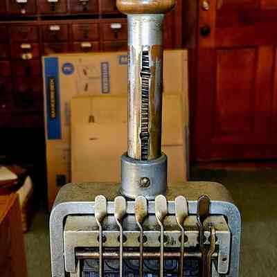 Stamp, Front View: The image depicts a vintage metal numbering stamp with a large wooden handle, designed to change numbers by adjusting small metal levers at the top; it features embossed numbers aligned in a rectangular frame, suggesting it was used for printing sequential numbers on paper or other materials. The background includes dark wooden cabinetry with numerous small drawers, indicative of an older office or library setting, alongside some books on a high shelf and cardboard boxes marked "medium" in the lower section, adding to the overall retro and utilitarian atmosphere conveyed by the scene.