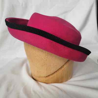A vibrant pink hat with a black band rests on a wooden stand against a white background.