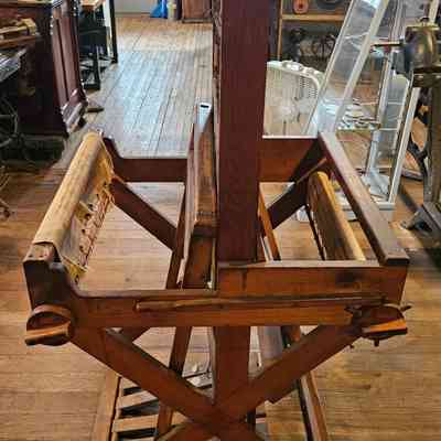 Caroline Stevens Rogers Loom, Right Side.: The image depicts a wooden loom, prominently placed in a historical setting, surrounded by vintage items and artifacts on display, including wooden cabinets, a typewriter, industrial machinery parts, and framed documents on the wall, suggesting an air of antiquity and craftsmanship; the floor is wooden, contributing to the overall rustic atmosphere, while natural light streams in from a window, casting soft shadows and highlighting the textures of the wooden surfaces and the machinery, evoking a sense of nostalgia for a bygone era of manual labor and artisanal skills.