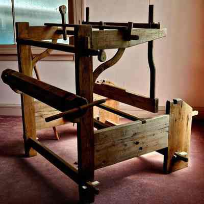 Assembled, but not yet conserved.: The image features a vintage wooden loom, set against the backdrop of a soft-lit room with a faded carpet, creating a nostalgic ambiance that highlights the craftsmanship of a bygone era. The loom's robust wooden frame, intricately constructed with various levers and supports, showcases the timeless design and mechanical ingenuity of traditional weaving equipment. Its rustic appearance is accentuated by the natural wood grain and aged patina, suggesting years of extensive use. Sunlight filters through a nearby window, casting gentle shadows that emphasize the loom's structural details and texture, while the simplicity of the surroundings, including a plain wall and muted floor, underscores the loom as the focal point, inviting the viewer to appreciate the beauty and history encapsulated in this piece of craftsmanship.