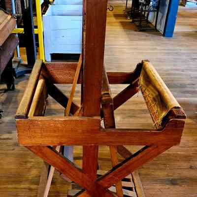 Caroline Stevens Rogers Loom, Left Side.: The image shows a wooden loom standing on a wooden floor in what appears to be a historical or antique setting, possibly a museum or workshop. The loom has a sturdy, X-shaped frame with two side supports made of brown wood, and a fabric is partially attached to it. Engraved on one of the wooden beams is the inscription "Property of Mrs. Horatio Rogers," suggesting ownership or dedication. Behind the loom, various other vintage items and furniture are visible, including a white crate, a sewing machine, and other tools, creating an atmosphere that evokes a sense of history and craftsmanship.