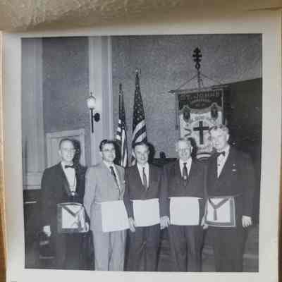 A square black and white photograph with a white deckled border. Five men wearing suits and white aprons.