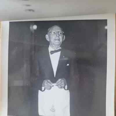 A square black and white photograph with a white deckled border. A man wearing a suit and a white apron with the Masonic compass on it.