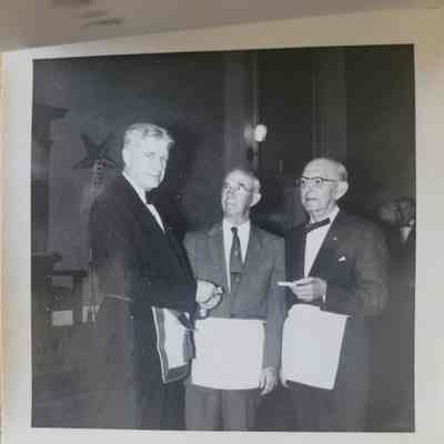 A square black and white photograph with a white deckled border. Three men wearing suits and white aprons.