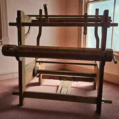 Assembled, but not yet conserved.: The image shows a large, wooden loom situated in a corner of a room with soft, natural light streaming in through a window, casting gentle shadows on the floor. The loom, made from solid wood, features various components such as beams, pegs, and levers that are essential for weaving textiles. It stands on a carpeted floor, against walls painted in a light color that enhances the serene and historic ambiance of the workspace. The simplicity and craftsmanship of the loom reflect an era when such tools were a central part of household industry, providing insight into traditional textile production methods.