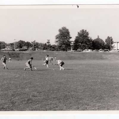 Women's field hockey match on Finnesey Field with Field House/Sports Complex in background.