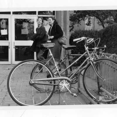 Two women students entering Bellarmine (?) with two bicycles parked in bicycle rack.
