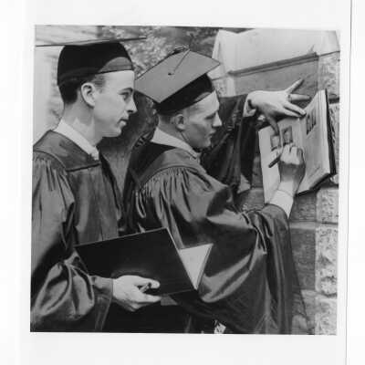 Two students dressed in cap and gowns signing yearbooks at graduation/commencement ceremony?