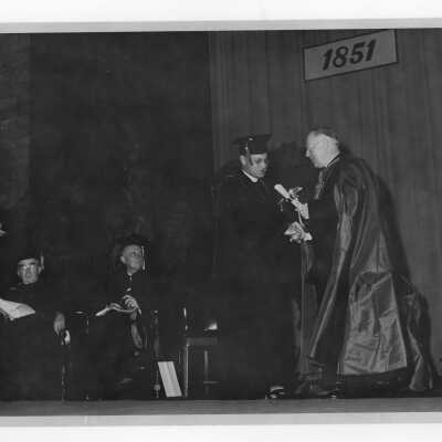 Edward G. Jacklin, S.J., conferring honorary degree on Bishop Thomas J. Feeney, S.J., during academic convocation, October 28, 1951, as part of centennial celebration. Joseph Hogan, S.J., and Thomas K. Finletter also on podium observing.