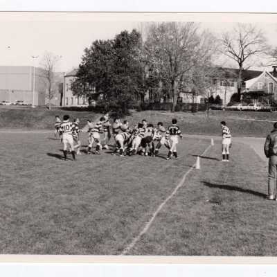Men's rugby game on Finnesey Field with Field House/Sports Complex in background.