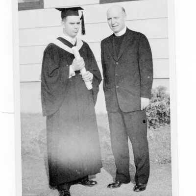 Graduated student standing with priest in front of the cardboard college after 1955 graduation/commencement cerermony.