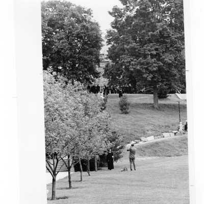 Graduate posing for family photograph on Finnesey Field during 1968 graduation/commencement. Other graduates are on the hill in the background.
