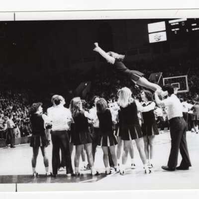 Men and women cheerleaders in circle catching cheerleader during basketball game.