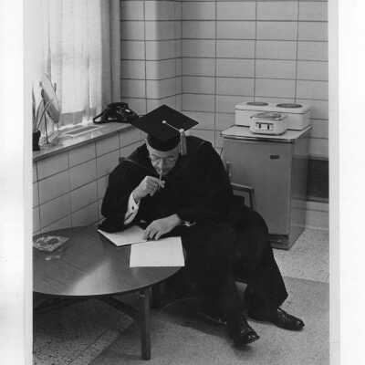 Paul C. Reinert, S.J., sitting in chair writing and wearing cap and gown during inauguration of Terrence Toland as president of Saint Joseph's College?