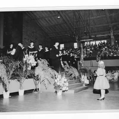 Edward G. Jacklin, S.J., awarding diplomas to graduates during 1956 graduation/commencement ceremony with young girl getting ready to take photograph.
