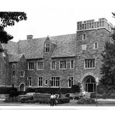 Exterior view of front of Barbelin building from City Avenue with two cars in driveway and four individuals unpacking.