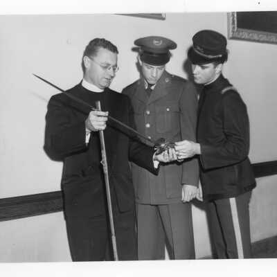 Edward G. Jacklin, S.J. with Tom Duffy in SJC cadet corps uniform and Bill Berry in ROTC uniform during centennial celebration event held October 21-28, 1951. The three individuals are looking at sword and scabbard (on loan from Breen family)