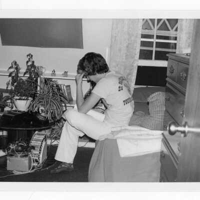 Student in dormitory room talking on rotary telephone on table with tape recorder and tray of cassettes nearby.