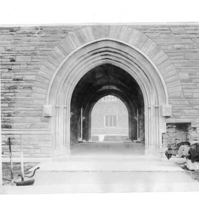 Exterior view of Barbelin archway near end of construction with stone masons' coats on bench and note on stone for gargoyle,"Don't ship, carve".