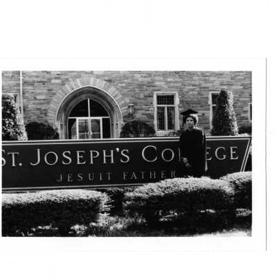 Student wearing cap and gown standing near Saint Joseph's College sign in front of Barbelin building.