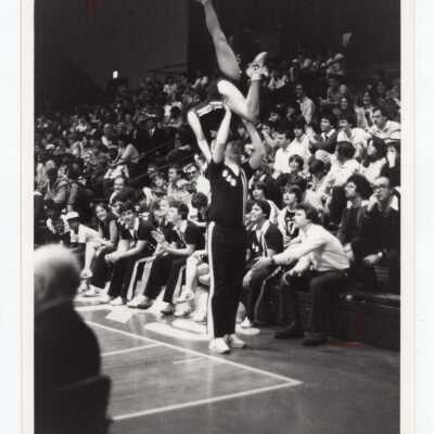 Two cheerleaders performing acrobatic routine and others sitting in bench during basketball game.