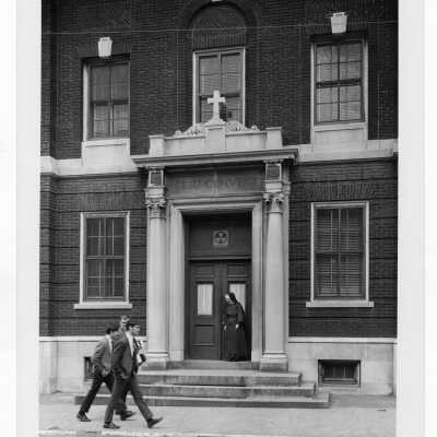Students wearing coat and tie walking in front of Gesu Convent in Philadelphia with nun standing at doorway.