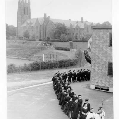 Students processing by Cardboard College and Veteran's Barracks as part of 1957 graduation/commencement ceremonies. Barbelin Hall and Finnesey Field are in background.