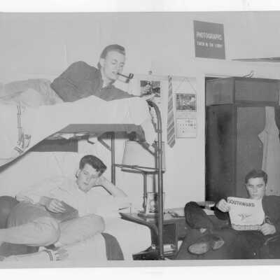 Three student residents of Barry Hall in dormitory room with bunk beds, desk, and closet and one student smoking pipe.