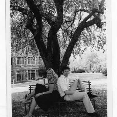 Two students sitting on park bench under tree near Barbelin building in spring.