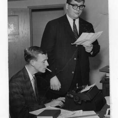 Two student staff members of The Hawk newspaper, Thomas Donohue and Francis Hargedon, reading copy and sitting at typewriter and smoking cigarette.