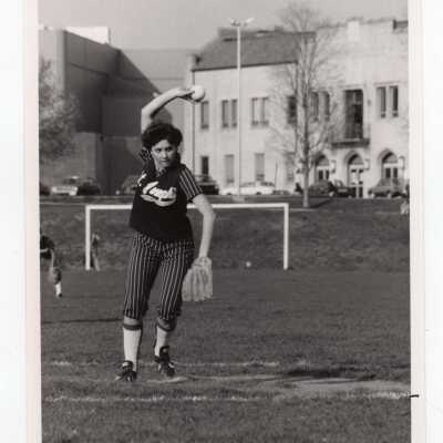 Women's softball team pitcher pitching in game against Lehigh University with Field House in background.
