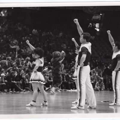 Men and women cheerleaders at basketball game with tiger mascot in background.