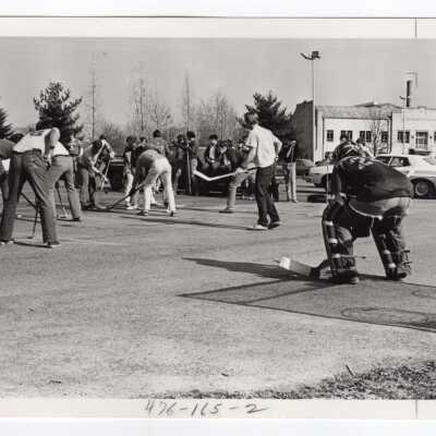 Men's ice hockey team practicing in parking lot with Field House in background.