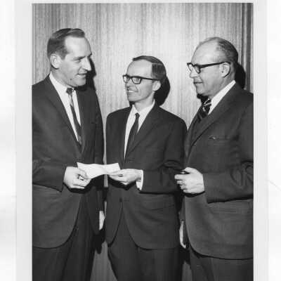 Harold S. Ricker (Food Marketing), one sitting at desk and the other with Ricker receiving check from two unidentified individuals.