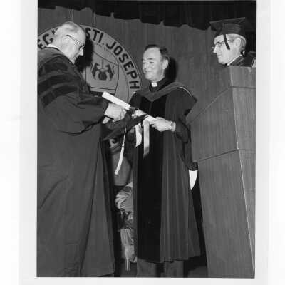Terrence Toland, S.J., conferring honorary degree to Harry Boyer (?) with Michael J. Smith, S.J. watching during convocation on December 17, 1975.
