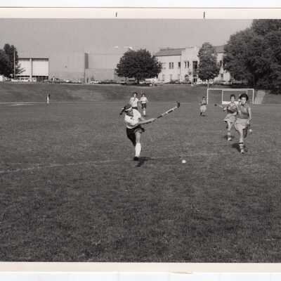 Women's field hockey match on Finnesey Field with Field House/Sports Complex in background.
