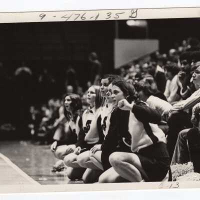 Women cheerleaders kneeling and watching basketball game.