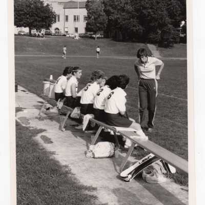 Women's field hockey players on bench during match with Field House in background.