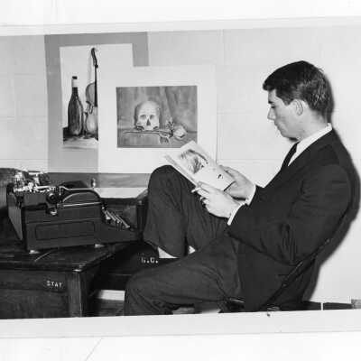 Student studying in dormitory room wearing coat and tie, smoking cigarette, with typewriter on desk.