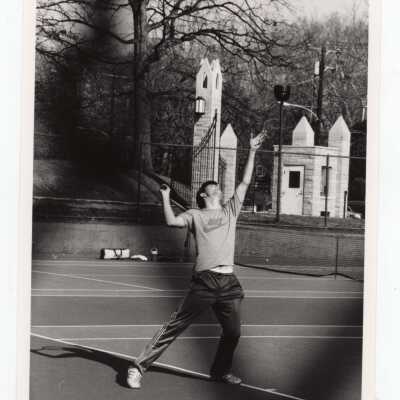 Men's tennis team member practicing with guard's kiosk and entrance gate in background.