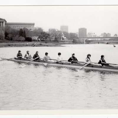 Women's crew/rowing team practicing on Schuylkill River with Philadelphia Museum of Art and city of Philadelphia skyline in background.
