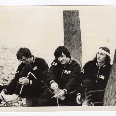 Three members of men's track team sitting on ground taking off shoes and wearing warm-up suits.