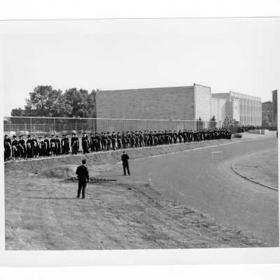 Students in cap and gown, including women, processing in front Villiger Hall toward Field House during 1966 graduation/commencement.