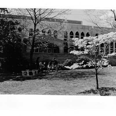Exterior front view of Drexel Library in spring with students and Terrence Toland, S.J. (?) sitting on bench in front of wall talking.