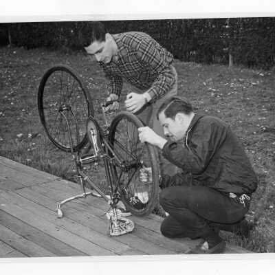 Two students preparing for bicycle race on wooden track near Field House during Junior Week activities.