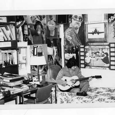 Male student in dormitory room playing guitar with walls decorated with posters.