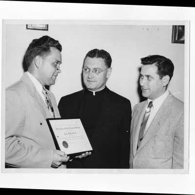 John J. Kraft holding alumni award certificate with Edward G. Jacklin, S.J., and William Ariano watching, c. 1953.